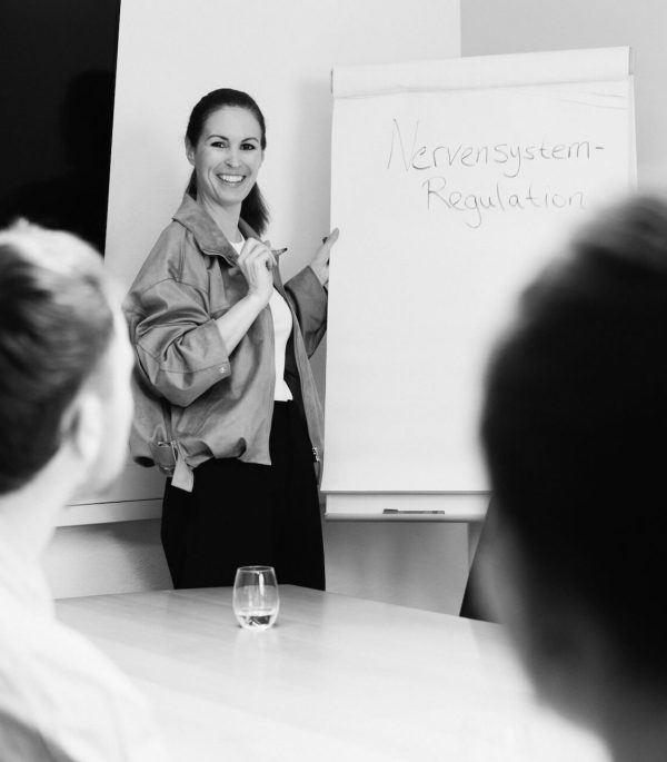 Schwarz-Weiß-Aufnahme von Christine Cottogni lächelnd vor einem Whiteboard während eines Workshops für Unternehmen zur Nervensystemregulation am Arbeitsplatz